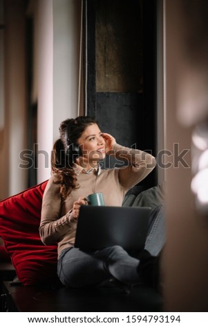 Beautiful young casual woman listening music with headphones near window at cafe restaurant or co working space. Businesswoman working on her laptop and enjoying music.