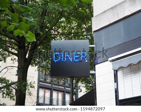 A lighted neon sign reads DINER attached to the corner of an old downtown building on a tree lined street. 