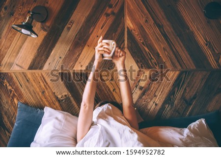 Lazy female lying under the white blanket on the  linen bed and holding the fresh coffee cup in the early morning. Lazy day off and "coffee in bed"concept image.