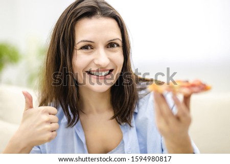 Pleased woman is smiles widely, on light background. Woman is showing her thumb finger up, holding a slice of yummy pizza. Close up.
