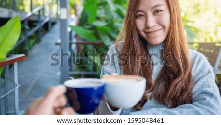 Close up image of a beautiful asian woman clinking coffee mugs with friend in cafe