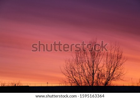 Colorful sunset landscape picture. Red clouds rises above rooftop and trees without leaves in autumn. Dramatic sky.