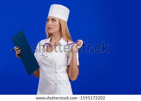 young woman doctor nurse in a white coat and hat with a folder for records tablet in hands posing on a blue background