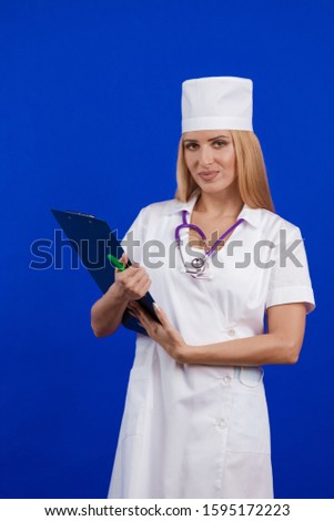 young woman doctor nurse in a white coat and hat with a folder for records tablet in hands posing on a blue background