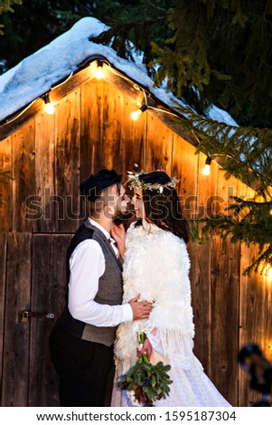 Newlywed couple, bride and groom on the background of an old wooden house and retro garland. Rustic winter wedding. Wedding ceremony at night. Unusual decorations, candles, light bulbs in the evening.