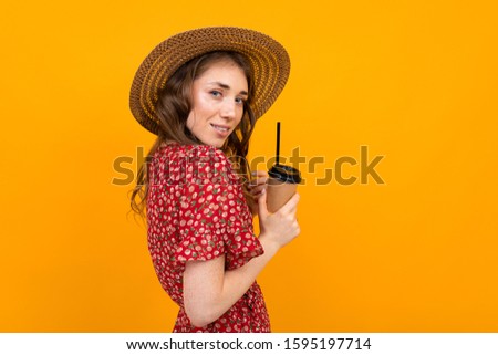 Studio portrait of a charming stylish smiling girl in a hat and a red dress with a paper cup on an isolated orange background