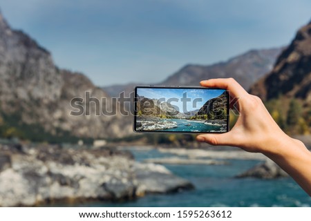Female hand with smartphone close-up. Taking photo on smartphone while traveling. Delightful mountain landscape, stormy river, rocks and blue sky.