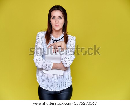 The concept of an office worker, teacher, manager. Portrait of a pretty brunette girl in a white business blouse smiling, talking to the camera on a yellow background with a folder in her hands.