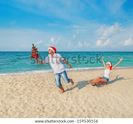 Young couple running at sea beach in red santa hats with color sled and decorated christmas tree - holiday concept