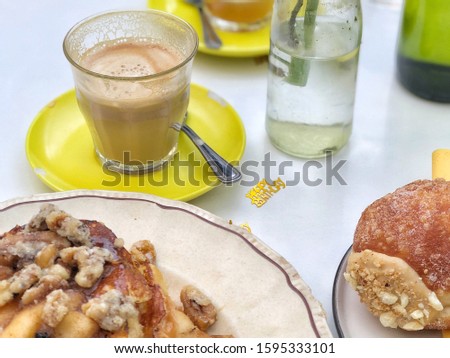White table with pancakes and donuts sweets, coffee drink, flower vase and happy birthday confetti. Coffee is on a yellow plate with spoon. It is bright