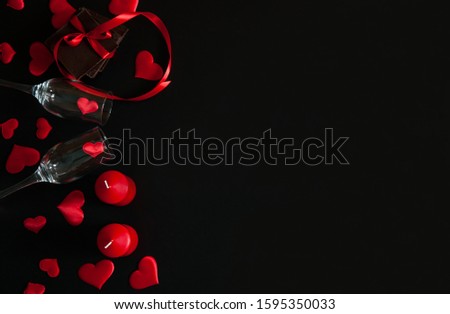 The Concept Of Valentine's Day. The border is made of two narrow empty glasses, chocolate tied with a red ribbon, candles and red hearts on a black background. Free space for your text.