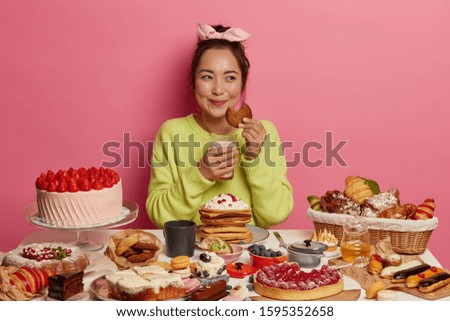 Sweet life, unhealthy eating concept. Cheerful Asian woman drinks milk with cookies, has good mood, surrounded with confectionery, gets more calories, being sweet tooth, isolated on pink wall. Royalty-Free Stock Photo #1595352658