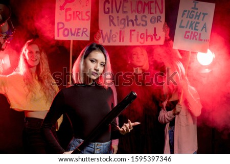 portrait of caucasian girl standing with bat promoting feminism isolated over smoky space in neon lights, group of feminists girls in the background
