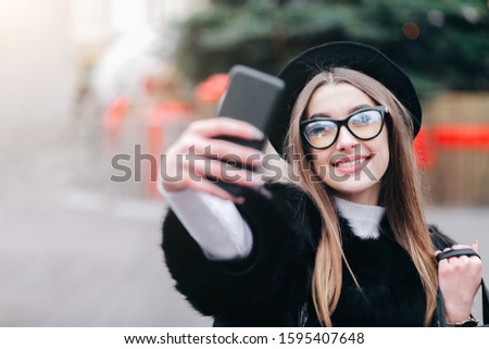 Pretty girl with shopping bags in hands makes a selfie on the background of the lights of a Christmas tree. Beautyful lady after Christmas shopping walks around the city and making selfie