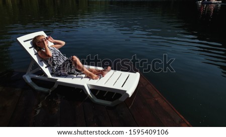 Kid girl lie on a sunbed in sunglasses and a boho silk shawl. Child rest on a flood wood underwater pier. The pavement is covered with water in the lake. In the background are mountain and a forest.