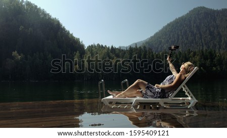 Woman shooting on handheld film gimbal stabilization for smartphone. Girl rest, relax, lie sunbed on pier in sunglasses, make selfie. Lady blogger broadcast video blogging vlogging. Take photo video.