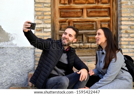 Portrait of excited adult couple man and woman taking selfie photo on cellphone and laughing sitting on the sidewalk