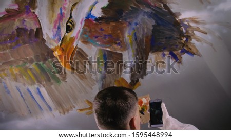 Artist designer draws an eagle on wall. Craftsman decorator paints picture with acrylic oil color looking at sketch in phone. Painter painter dressed in paint coat. Indoor. Dark magic cinematic look.