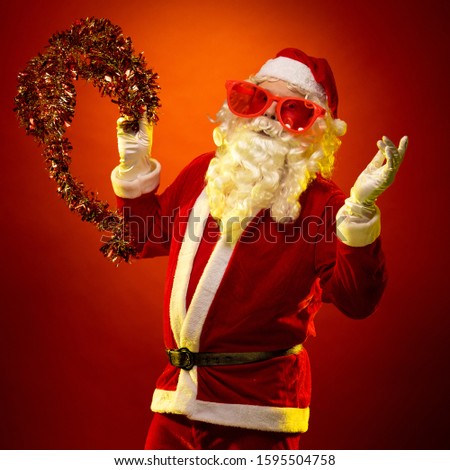 Santa Claus in large pink glasses and with a red garland of tinsel in his hands dancing and posing on a dark red background