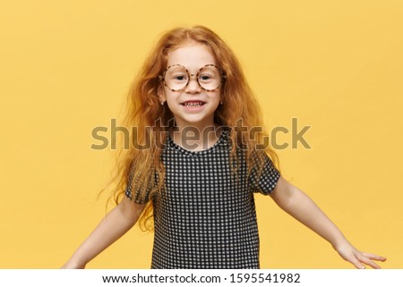 Funny little girl with long loose red hair jumping expressing true positive emotions smiling broadly wearing large round spectacles. Picture of cute cheerful child having fun posing isolated