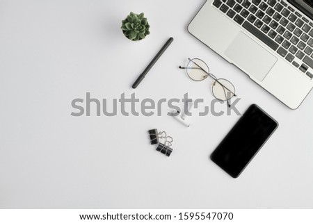 Wireless earphones, eyeglasses, pen, clips, smartphone, small domestic plant and laptop keypad on white desk