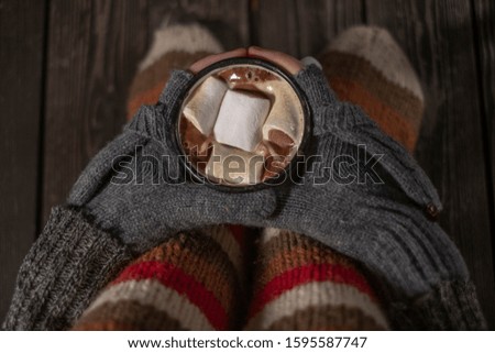 Female hands in gloves with fingerless mittens bask on tasty marshmallows in an old white iron retro mug with aromatic coffee or cocoa, against the background of legs in knitted woolen socks on a wood