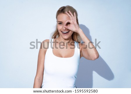 Portrait of a happy young blonde girl showing ok gesture over her eyes and winking isolated over grey background.