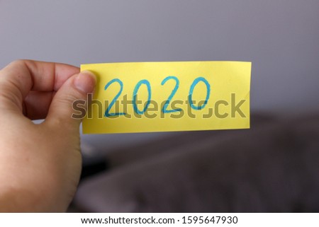 hand holding yellow paper with 2020 handwritten in pencil with blurred background