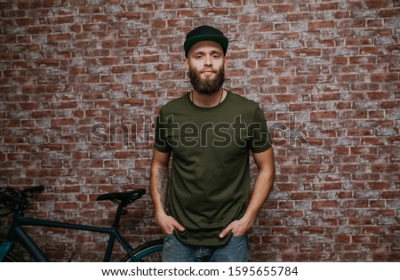 City portrait of handsome hipster guy with beard wearing a blank green military t-shirt and blank black cap standing on a brick wall background. Empty space for your logo or design. Mockup for print.