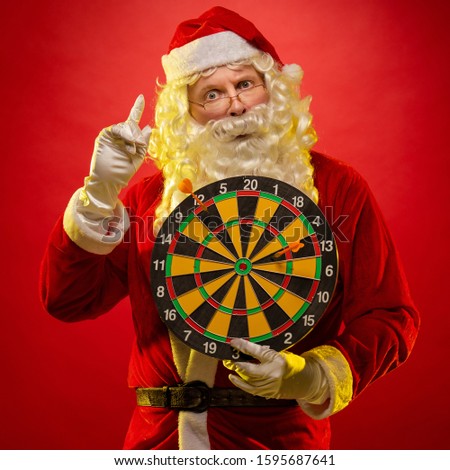 Santa Claus holds a darts target in his hands and poses on a dark red background