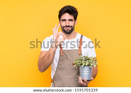 Gardener man with beard over isolated yellow background showing an ok sign with fingers