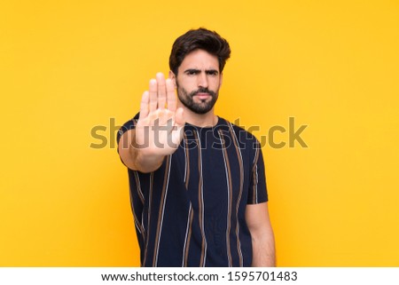 Young handsome man with beard over isolated yellow background making stop gesture with her hand