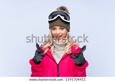 Skier teenager girl with snowboarding glasses over isolated blue background points finger at you