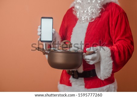 Father Santa using cell phone showing holding cooking pot. Presents time of joy celebration in traditional costume. Internet browsing copyspace display delivering shop idea solution. Closeup photo