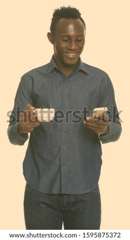 Young happy black African man smiling and holding coffee cup while using mobile phone