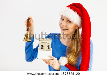 Happy positive young teenage woman wearing Santa Claus christmas hat holding calendar with 24 december date and bell