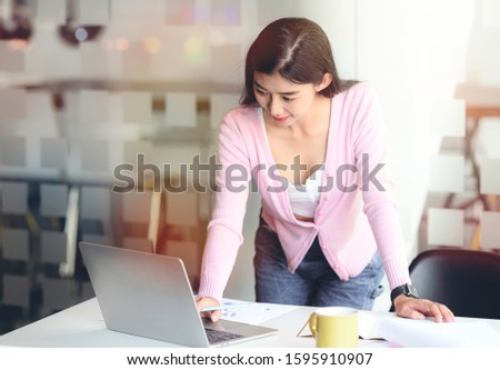 Young attractive businesswoman in casual wear standing at office desk and working with laptop with happiness.