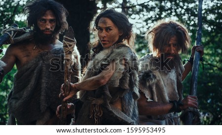 Female Leader and Two Primeval Cavemen Warriors Threat Enemy with Stone Tipped Spear, Scream, Defending Their Cave and Territory in the Prehistoric Times. Neanderthals / Homo Sapiens Tribe Royalty-Free Stock Photo #1595953495