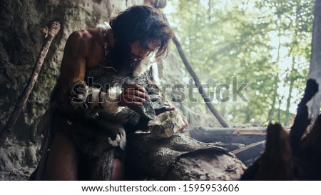Primeval Caveman Wearing Animal Skin Hits Rock with Sharp Stone and Makes First Primitive Tool for Hunting Animal Prey or to Handle Hides. Neanderthal Using Handax. Dawn of Human Civilization Royalty-Free Stock Photo #1595953606