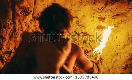 Primeval Caveman Wearing Animal Skin Standing in His Cave At Night, Holding Torch with Fire Looking at Drawings on the Walls at Night. Cave Art with Petroglyphs, Rock Paintings. Back View Royalty-Free Stock Photo #1595983408