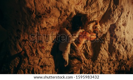 Primitive Prehistoric Neanderthal Wearing Animal Skin Draws Animals and Abstracts on the Walls at Night. Creating First Cave Art with Petroglyphs, Rock Paintings Illuminated by Fire. Back View
