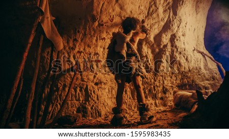 Back View of Primitive Prehistoric Neanderthal Wearing Animal Skin Draws Animals and Abstracts on the Walls at Night. Creating First Cave Art with Petroglyphs, Rock Paintings Illuminated by Fire