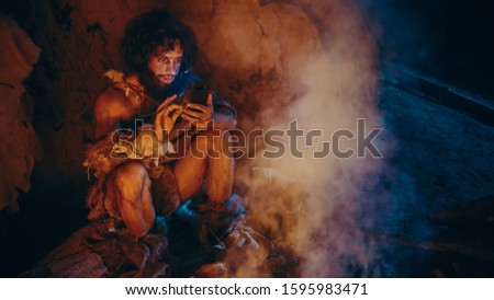 Tribe of Prehistoric, Primitive Hunter Gatherer Wearing Animal Skin Uses Smartphone in a Cave at Night. Neanderthal / Homo Sapiens Male Browsing Internet on Mobile Phone, Watches Videos Royalty-Free Stock Photo #1595983471
