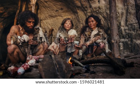 Tribe of Prehistoric Hunter-Gatherers Wearing Animal Skins Live in a Cave at Night. Neanderthal or Homo Sapiens Family Trying to Get Warm at the Bonfire, Holding Hands over Fire, Cooking Food Royalty-Free Stock Photo #1596021481