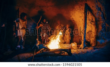 Tribe of Prehistoric Hunter-Gatherers Wearing Animal Skins Stand Around Bonfire Outside of Cave at Night. Portrait of Neanderthal / Homo Sapiens Family Doing Pagan Religion Ritual Near Fire Royalty-Free Stock Photo #1596021505
