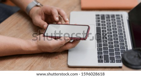 Cropped shot of young man using blank screen smartphone while working on his project with laptop computer 