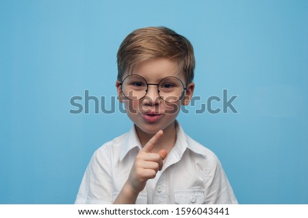 Funny little caucasian boy in glasses shows a sign of silence bowing his finger over a card on a blue background. Concept of personal offers and promotions in children's stores. Advertising space