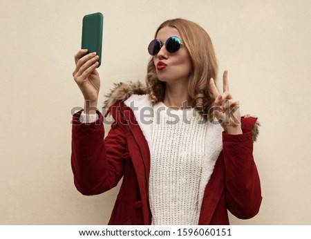 Portrait of beautiful young woman taking selfie with smartphone blowing her lips sends kiss wearing red jacket with fur hood