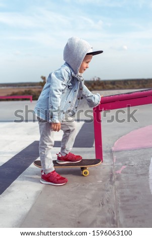 Little boy 3-5 years old, learning to ride a skateboard, autumn day, casual warm clothes. Denim sweater with a hood. Driving lessons, first experience, outdoor sports