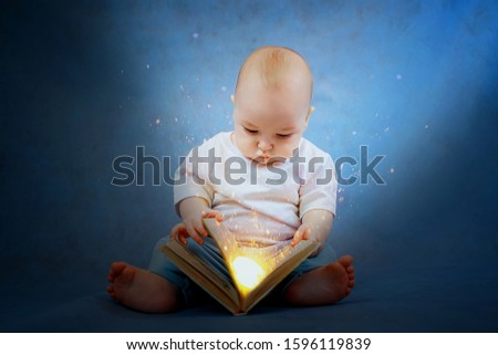 A cute little kid in light clothes with curiosity leafs through a book from which a bright light pours on him. Fairytale picture. Education concept.